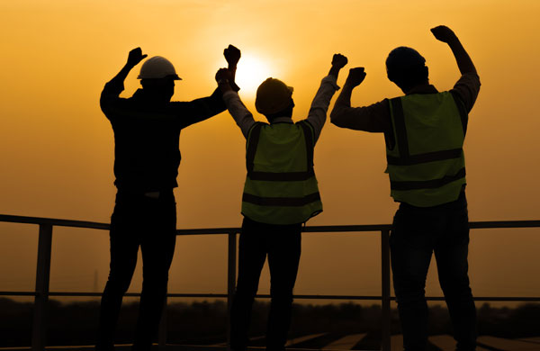 Three workers with hardhats cheering, facing sunset. In sillouette.